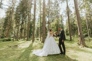 Fashionable groom and cute bride in white dress with train and crown on head walking happily in park, garden, forest outdoors. Wedding photography, portrait of smiling newlyweds. photo