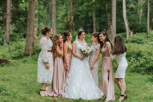 A group of beautiful girls with a bride in identical dresses are smiling, celebrating and having fun together against the background of nature and tall trees. Girls party photo