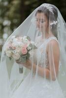 the bride in a wedding dress with a long train and a veil holds a wedding bouquet of roses, under the veil photo