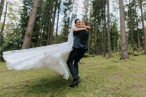 de moda novio y linda novia en blanco vestir con largo tren y corona en cabeza, novio dando vueltas con novia en brazos, jardín, bosque al aire libre. Boda fotografía, retrato de sonriente recién casados. foto