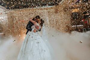 Gorgeous bride and stylish groom dancing under golden confetti at wedding reception. Happy wedding couple performing first dance in restaurant. Romantic moments photo