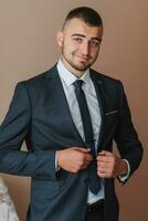 Confident elegant handsome guy standing at home and buttoning his jacket looking at the camera in a nice dark blue suit photo