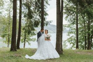A wedding couple is enjoying the best day of their lives against the backdrop of a lake and tall trees. The groom hugs the bride from behind photo