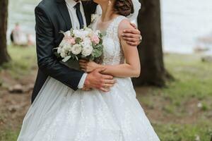 A wedding couple is enjoying the best day of their lives against the backdrop of a lake and tall trees. The groom hugs the bride. photo