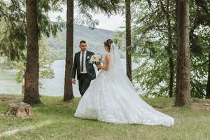 A wedding couple is enjoying the best day of their lives against the backdrop of a lake and tall trees. The bride and groom are walking in nature. He leads her forward by the hand photo