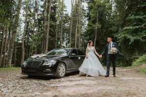 Front view of a married bride and groom wearing festive clothes standing against a black car on their wedding day photo