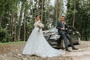 Front view of a married bride and groom wearing festive clothes standing against a black car on their wedding day photo