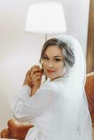 A beautiful bride in a robe and lush veil, with beautiful hair and makeup, poses in her room. Dressing up and preparing for the wedding ceremony photo