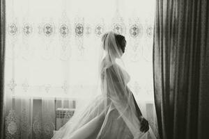 Portrait of the bride. The bride is dressed in an elegant dress, posing by the window wearing earrings. Tiara and jewelry. Open bust. black and white photo