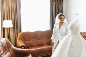 A beautiful bride in a robe with beautiful hair and makeup, standing next to her dress on a mannequin. Dressing up and preparing for the wedding ceremony photo