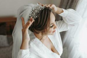 A beautiful bride in a robe and lush veil, with beautiful hair and makeup, poses while adjusting her tiara. Dressing up and preparing for the wedding ceremony photo