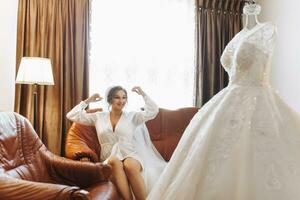 A beautiful bride in a robe with beautiful hair and makeup, sitting on a luxurious sofa, next to her dress on a mannequin. Dressing up and preparing for the wedding ceremony photo