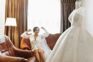 un hermosa novia en un túnica con hermosa pelo y constituir, sentado en un lujoso sofá, siguiente a su vestir en un maniquí. vendaje arriba y preparando para el Boda ceremonia foto