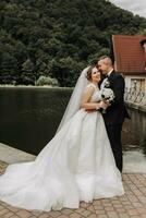 A brunette bride in a long dress and a groom in a classic suit stand embracing on a bridge near a lake with a castle in the background. photo