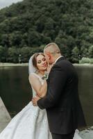 A brunette bride in a long dress and a groom in a classic suit stand embracing on a bridge near a lake with a castle in the background. photo