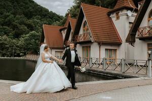 un morena novia en un largo vestir y un novio en un clásico traje son caminando en un puente cerca un lago en contra el antecedentes de un castillo. un caminar en naturaleza. Boda día foto