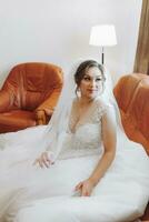 Beautiful bride in a white dress with beautiful hair and makeup sitting on a luxurious sofa. Dressing up and preparing for the wedding ceremony photo