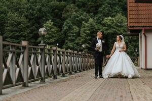 A brunette bride in a long dress and a groom in a classic suit are walking on a bridge near a lake against the background of a castle. a walk in nature. Wedding day photo