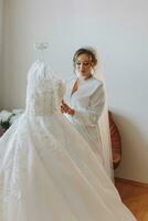 A beautiful bride in a robe with beautiful hair and makeup, standing next to her dress on a mannequin. Dressing up and preparing for the wedding ceremony photo