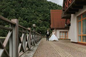 A brunette bride in a long dress and a groom in a classic suit are walking on a bridge near a lake against the background of a castle. a walk in nature. Wedding day photo