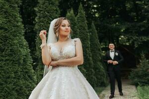 Wedding portrait. The bride in an elegant dress stands in front of the groom in a classic suit, against the background of green trees. Gentle touch. Summer wedding. A walk in nature photo