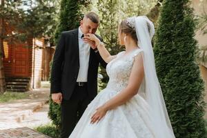Wedding portrait. The groom kisses the hand of the bride. The bride in an elegant dress, the groom in a classic suit against the background of green trees. Gentle touch. A walk in nature photo