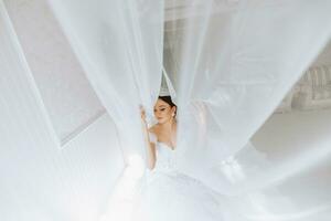 preparación para el boda. hermosa joven novia en blanco lujoso Boda vestido, tiara en cabeza, largo velo en real hotel habitación. lujo sonriente modelo foto