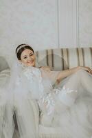 un hermosa niña con un Boda peinado y un tiara en su cabeza en un transparente túnica es preparando para un Boda en un hotel con un real interior. largo velo, natural luz, retrato foto