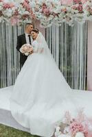 Attractive bride and groom at the ceremony on their wedding day with an arch made of pink and white flowers. Beautiful newlyweds, a young woman in a white dress with a long train, men in a black suit. photo