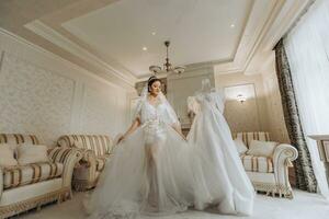 a beautiful girl with a wedding hairstyle in a transparent robe is preparing for a wedding in a hotel with a royal interior. The bride poses next to her wedding dress on a mannequin photo