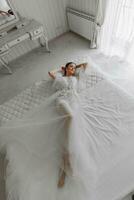 a beautiful girl with a wedding hairstyle and a tiara on her head in a transparent robe, lying on a bed in a hotel with a royal interior. Long veil, natural light, portrait photo