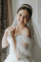 Preparation for the wedding. Beautiful young bride in white luxurious wedding dress, tiara on head, long veil in royal hotel room. Luxury smiling model photo