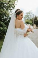 Rear view portrait of beautiful bride in white wedding dress with long train with modern hairstyle and veil walking in garden. Wedding concept photo