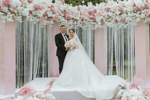 atractivo novia y novio a el ceremonia en su Boda día con un arco hecho de rosado y blanco flores hermosa recién casados, un joven mujer en un blanco vestir con un largo tren, hombres en un negro traje. foto