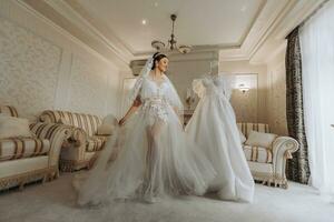 a beautiful girl with a wedding hairstyle in a transparent robe is preparing for a wedding in a hotel with a royal interior. The bride poses next to her wedding dress on a mannequin photo