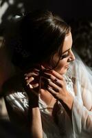 Morning of the bride before the wedding. A beautiful young woman with a long veil in a white robe on a dark background fastens an earring. Natural beauty and professional makeup. photo