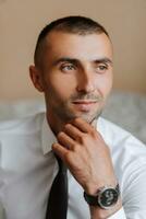 A stylish groom with a watch on his hand and a tie is sitting on a chair in an expensive hotel room. Groom's morning. The groom is getting ready in the morning before the wedding ceremony. photo