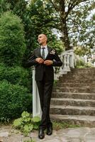 un hombre en un clásico negro traje y blanco camisa soportes y poses cerca el Roca pasos. un elegante mirar. de los hombres estilo. moda. negocio. foto