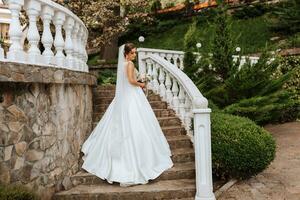 un hermosa morena novia en un blanco largo vestir con un ramo de flores de flores soportes en el escalera en contra el antecedentes de alto árboles, al aire libre en el parque. foto