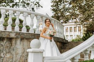 un hermosa morena novia en un blanco largo vestir con un ramo de flores de flores soportes en el escalera en contra el antecedentes de alto árboles, al aire libre en el parque. foto