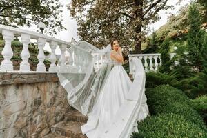 A beautiful brunette bride in a white long dress with a bouquet of flowers stands on the stairs against the background of tall trees, outdoors in the park. photo