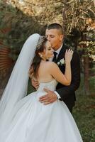 A stylish groom in a black suit and a cute bride in a white dress with a long veil are hugging in a park. Wedding portrait of smiling and happy newlyweds. photo