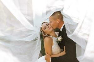 gorgeous happy luxurious brunette bride and elegant stylish groom stand under a veil and embrace tenderly photo