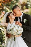 A stylish groom in a black suit and a cute bride in a white dress with a long veil are hugging in a park. Wedding portrait of smiling and happy newlyweds. photo