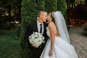 A stylish groom in a black suit and a cute bride in a white dress with a long veil are hugging in a park. Wedding portrait of smiling and happy newlyweds. photo