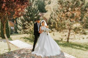 A stylish groom in a black suit and a cute bride in a white dress with a long veil are hugging in a park. Wedding portrait of smiling and happy newlyweds. photo