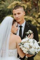 A stylish groom in a black suit and a cute bride in a white dress with a long veil are hugging in a park. Wedding portrait of smiling and happy newlyweds. photo