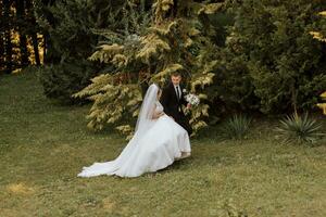 A stylish groom in a black suit and a cute bride in a white dress with a long veil are hugging and walking near green tall trees. Wedding portrait of smiling and happy newlyweds. photo