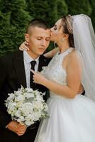 A stylish groom in a black suit and a cute bride in a white dress with a long veil are hugging in a park. Wedding portrait of smiling and happy newlyweds. photo