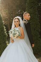 A stylish groom in a black suit and a cute bride in a white dress with a long veil are hugging in a park. Wedding portrait of smiling and happy newlyweds. photo